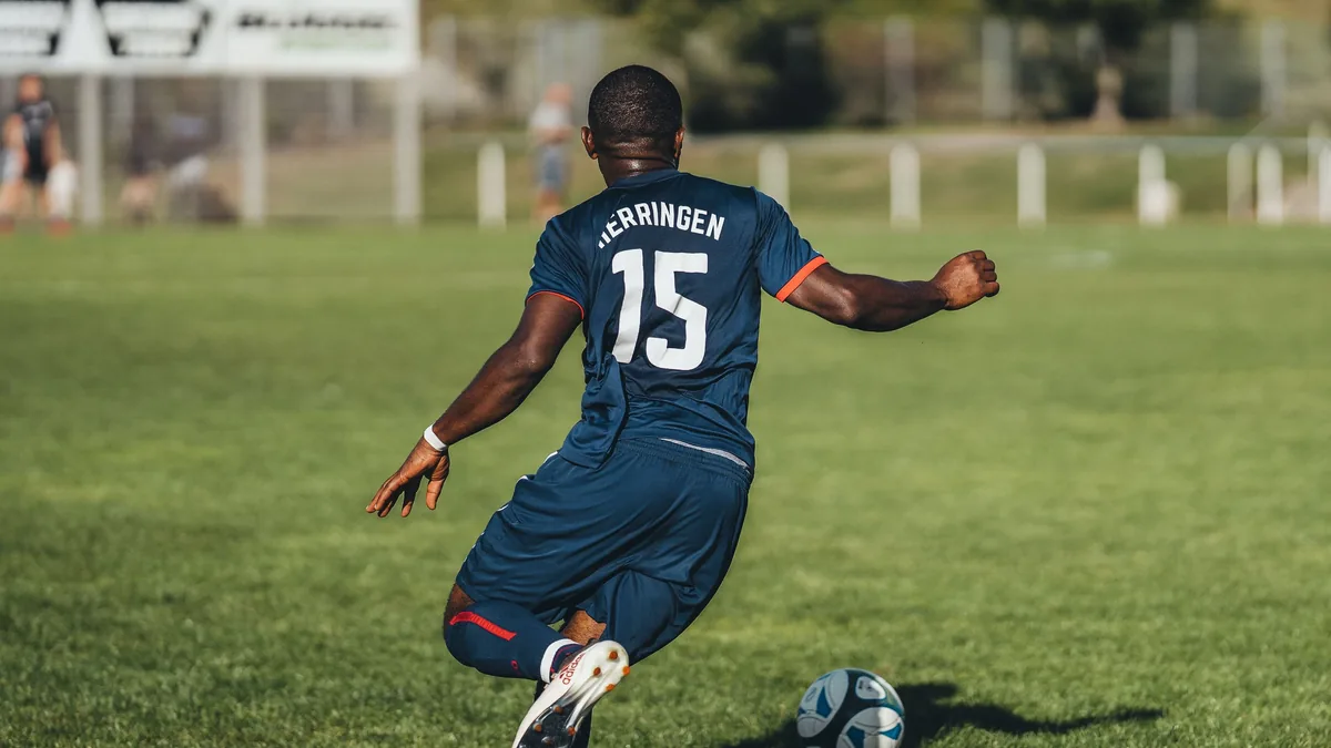 A soccer player in a blue uniform, with the number 15 on his back, kicking a soccer ball on a grassy field.